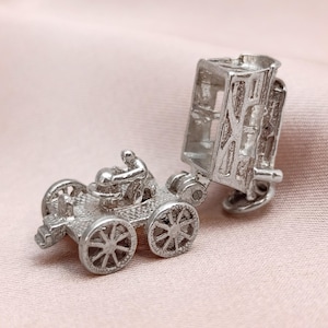 May include: A silver charm depicting a vintage car with an open carriage. The car features four spoked wheels and a driver figure. The carriage section is hinged, allowing it to open. This charm is likely a collectible or a piece of jewellery.