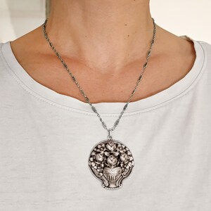 Pu&ograve; includere: Collana con pendente argentato con un design floreale dettagliato. Il pendente raffigura un vaso pieno di rose e altri fiori, circondato da un bordo decorativo. La collana ha una catena delicata.