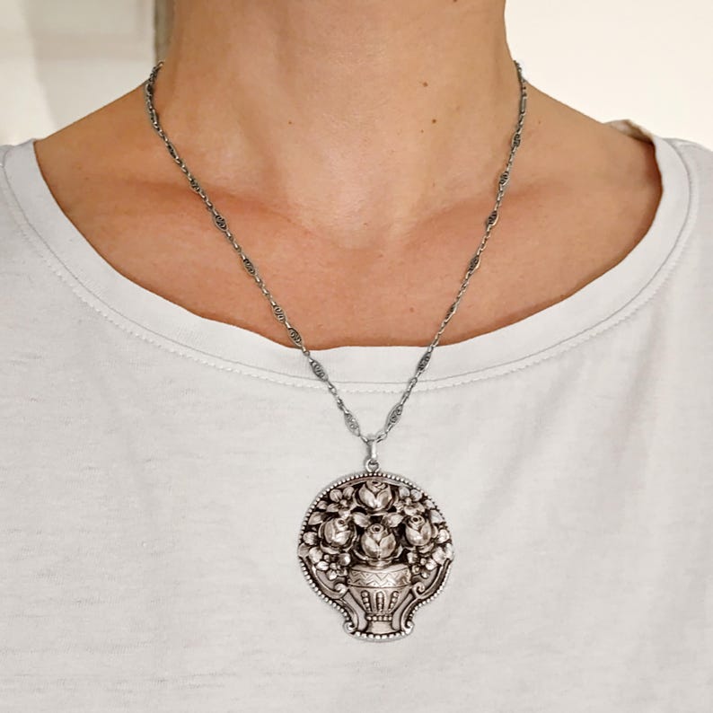 Pu&ograve; includere: Collana con pendente argentato con un design floreale dettagliato. Il pendente raffigura un vaso pieno di rose e altri fiori, circondato da un bordo decorativo. La collana ha una catena delicata.