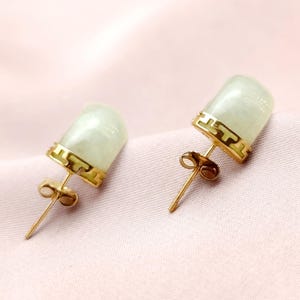 May include: A pair of stud earrings featuring pale green jade stones set in gold-colored metal. The earrings have a cylindrical shape with a decorative gold band at the top. The background is a soft pink fabric.