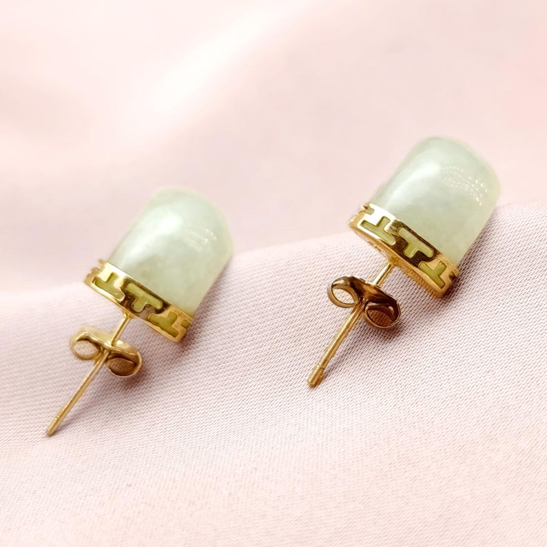 May include: A pair of stud earrings featuring pale green jade stones set in gold-colored metal. The earrings have a cylindrical shape with a decorative gold band at the top. The background is a soft pink fabric.