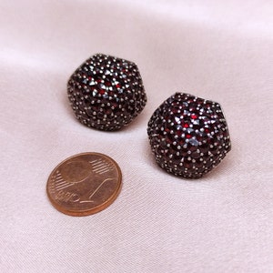 May include: A pair of silver earrings set with red garnet stones. The earrings are round and have a textured surface. The earrings are approximately the size of a UK one pence coin.