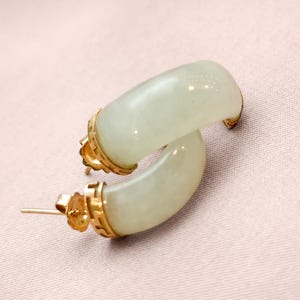 May include: A pair of jade hoop earrings with gold accents. The earrings have a curved, semi-circular design with a pale green hue. The gold settings add a touch of elegance to the jewelry.