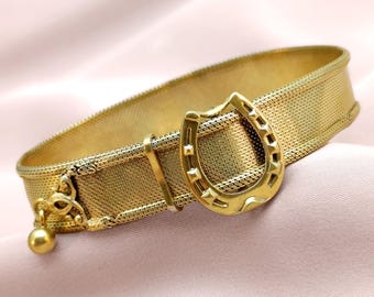 Puede incluir: Una pulsera dorada con una banda de malla y una hebilla en forma de herradura. La pulsera presenta un pequeño dije decorativo con una bola dorada colgante. El diseño es vintage y elegante.