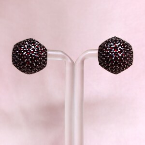 May include: A pair of silver earrings set with red garnet stones. The earrings are round and have a textured surface.