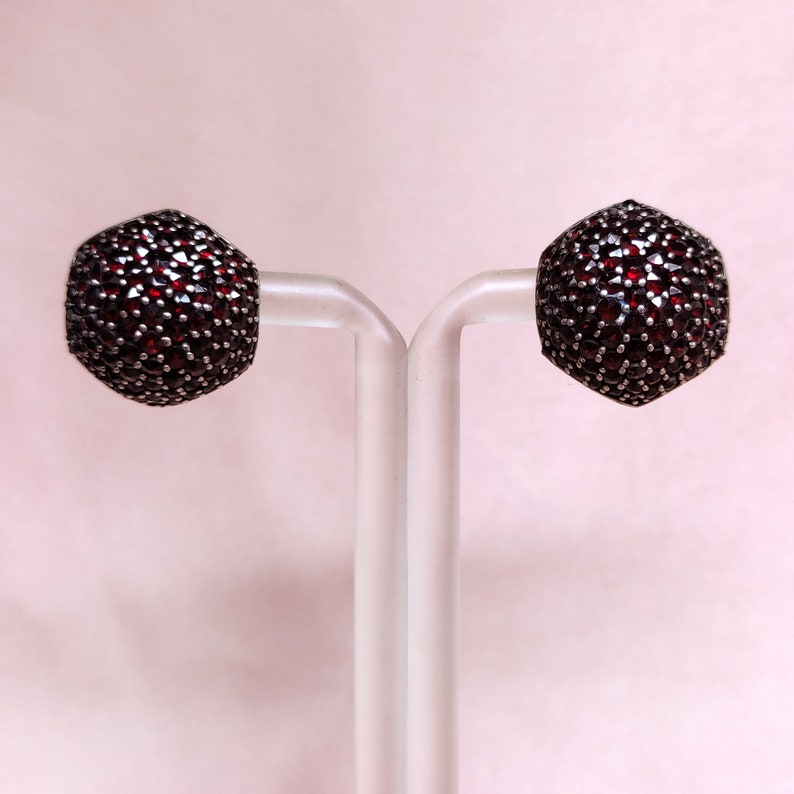 May include: A pair of silver earrings set with red garnet stones. The earrings are round and have a textured surface.