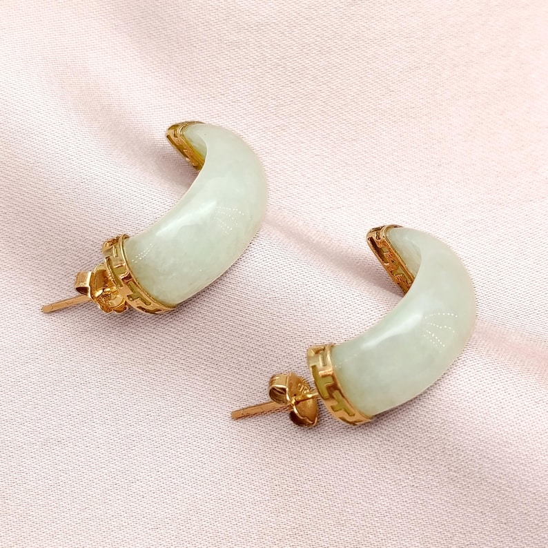 May include: A pair of crescent-shaped earrings. Each earring features a pale green jade stone set in a gold-colored metal frame. The earrings have a post-and-butterfly-clasp closure. The background is a light pink fabric.