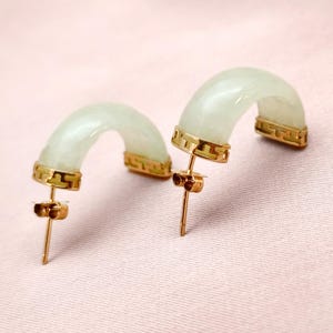 May include: A pair of jade hoop earrings with gold-colored accents. The earrings have a curved, semi-circular design with a pale green hue. The gold-colored metal frames the jade and features a geometric pattern. The earrings are displayed on a pink surface.
