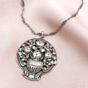 Pu&ograve; includere: Collana con pendente argentato con un design floreale dettagliato. Il pendente mostra un vaso traboccante di rose e altri fiori. La collana ha una catena delicata e una chiusura decorativa. Il pendente misura circa 5 cm di diametro.