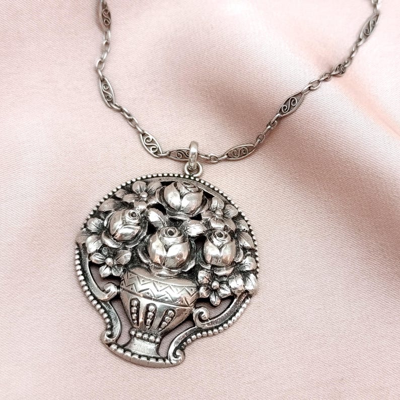 Pu&ograve; includere: Collana con pendente argentato con un design floreale dettagliato. Il pendente mostra un vaso traboccante di rose e altri fiori. La collana ha una catena delicata e una chiusura decorativa. Il pendente misura circa 5 cm di diametro.