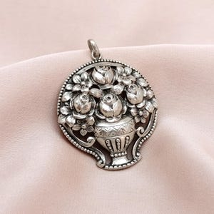 Pu&ograve; includere: Ciondolo color argento con un design floreale. Il ciondolo raffigura un vaso pieno di rose e altri fiori, circondato da un bordo di perline. Ha un anello per una catena.