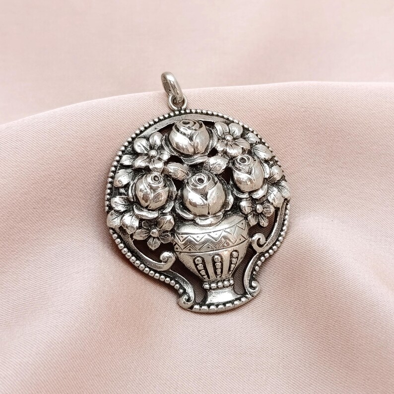 Pu&ograve; includere: Ciondolo color argento con un design floreale. Il ciondolo raffigura un vaso pieno di rose e altri fiori, circondato da un bordo di perline. Ha un anello per una catena.
