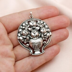 Pu&ograve; includere: Ciondolo argentato con design floreale. Il ciondolo raffigura un vaso pieno di rose e altri fiori, circondato da un bordo decorativo. Ha un anello nella parte superiore per una catena.