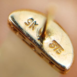 May include: Close-up of a gold earring back with the markings "9K" and "375" visible. The earring back is oval-shaped and has a post for insertion into the ear. The gold has a polished finish.