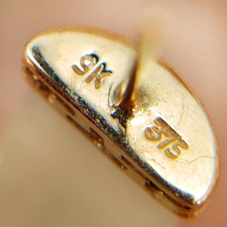 May include: Close-up of a gold earring back with the markings "9K" and "375" visible. The earring back is oval-shaped and has a post for insertion into the ear. The gold has a polished finish.