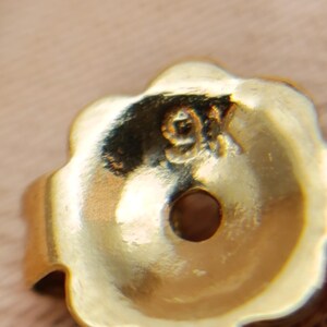 May include: Close-up of a gold-colored earring back with a flower-like shape. The earring back has a small hole in the center and the marking "9K" is visible. The background is a light tan color.