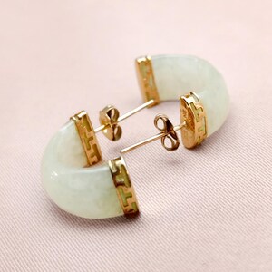 May include: A pair of jade earrings with gold accents. The earrings have a curved, half-moon shape with a pale green hue. The gold settings feature a decorative pattern. The earrings are displayed on a pink surface.