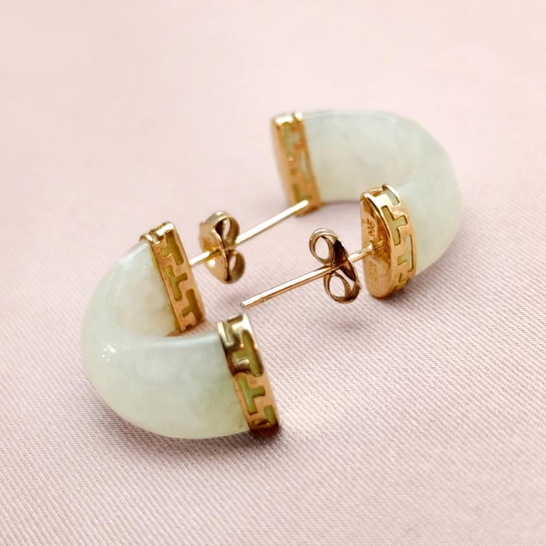 May include: A pair of jade earrings with gold accents. The earrings have a curved, half-moon shape with a pale green hue. The gold settings feature a decorative pattern. The earrings are displayed on a pink surface.