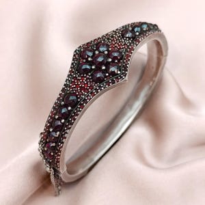 Könnte beinhalten: Ein silbernes Klapparmband mit geometrischem Design. Das Armband ist mit zahlreichen dunkelroten Edelsteinen verziert. Die Edelsteine sind in einem detaillierten Muster gesetzt, das einen Kontrast zum Silbermetall bildet.