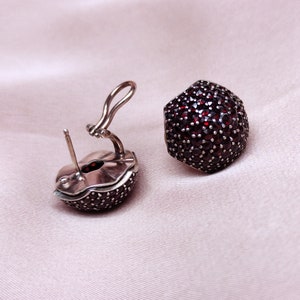 May include: A pair of silver earrings with a round, red gemstone design. The earrings are made of silver and have a hinged closure.