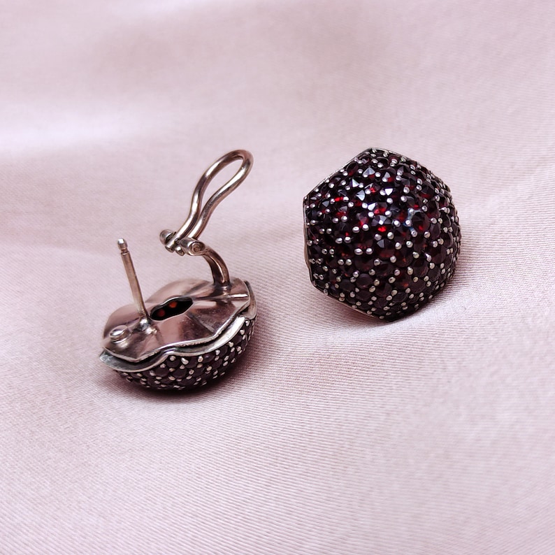 May include: A pair of silver earrings with a round, red gemstone design. The earrings are made of silver and have a hinged closure.