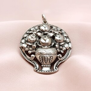 Pu&ograve; includere: Un ciondolo argentato raffigurante un vaso con un'arrangiamento floreale dettagliato. Il ciondolo presenta rose e altri fiori, con dettagli intricati sui bordi. Il ciondolo ha un anello nella parte superiore per una catena.