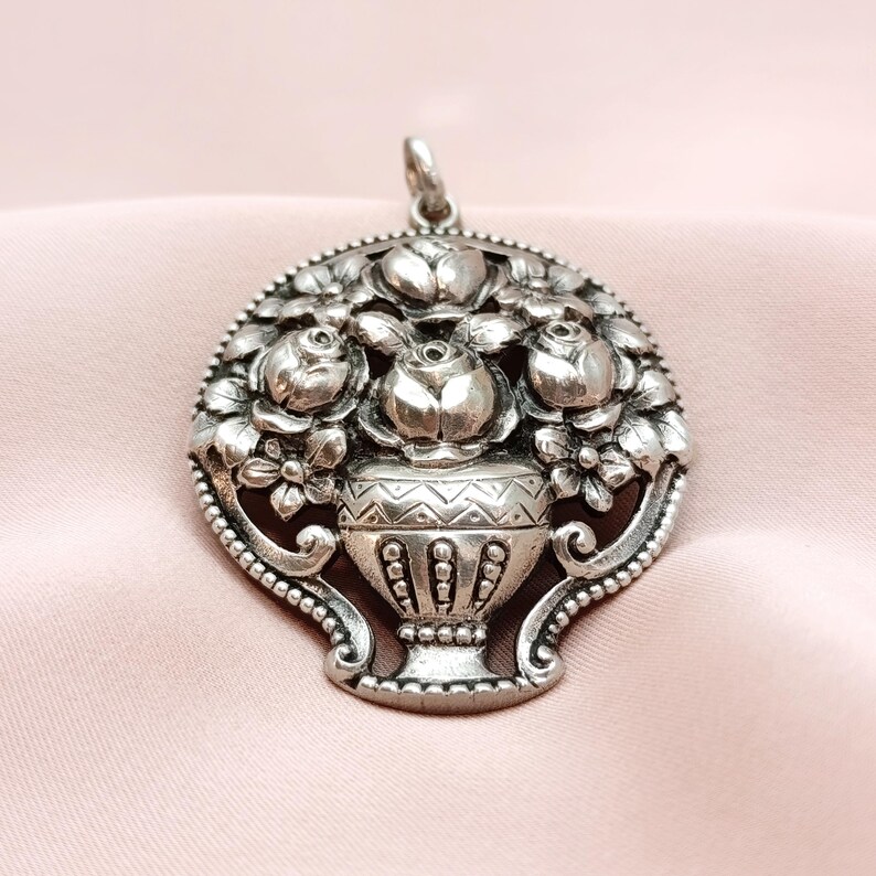 Pu&ograve; includere: Un ciondolo argentato raffigurante un vaso con un'arrangiamento floreale dettagliato. Il ciondolo presenta rose e altri fiori, con dettagli intricati sui bordi. Il ciondolo ha un anello nella parte superiore per una catena.