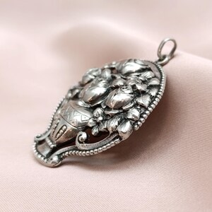 Pu&ograve; includere: Un ciondolo argentato di forma ovale, con un rilievo dettagliato di un vaso traboccante di fiori. Il ciondolo ha un bordo strutturato e un anello per una catena. Il design &egrave; intricato e ornato, con un'estetica vintage.