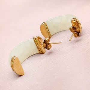 May include: A pair of gold-toned hoop earrings with a light green, curved jade center. The earrings have a post-and-clutch closure. The gold accents feature intricate detailing. The earrings are displayed on a pink surface.