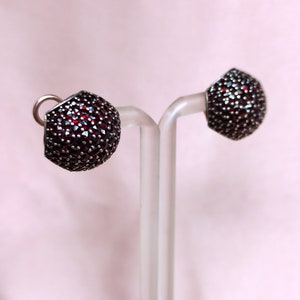 May include: A pair of silver earrings with red gemstone accents. The earrings are round and have a textured surface.