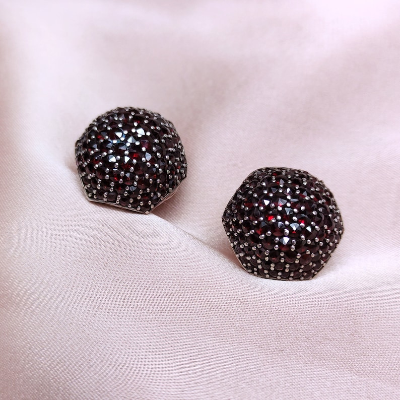 May include: A pair of silver earrings with a hexagonal shape, each earring is set with a cluster of small red gemstones.