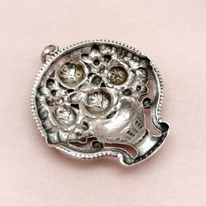 Pu&ograve; includere: Ciondolo argentato con design floreale. Il ciondolo raffigura un bouquet di rose in una forma a vaso, incorniciato da un bordo dettagliato e strutturato. Il design ha un'estetica vintage.