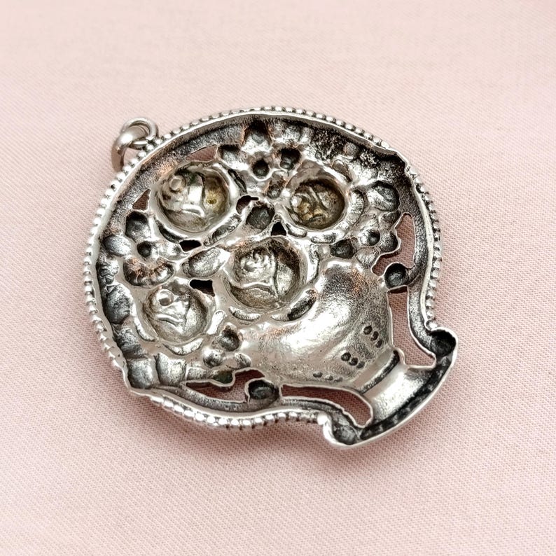 Pu&ograve; includere: Ciondolo argentato con design floreale. Il ciondolo raffigura un bouquet di rose in una forma a vaso, incorniciato da un bordo dettagliato e strutturato. Il design ha un'estetica vintage.