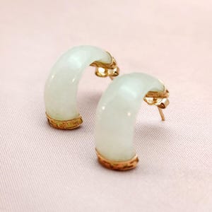 May include: A pair of jade hoop earrings with gold accents. The earrings have a curved, crescent shape with a pale green hue. The gold detailing is visible at the top and bottom of the jade hoops. The earrings are displayed on a pink surface.