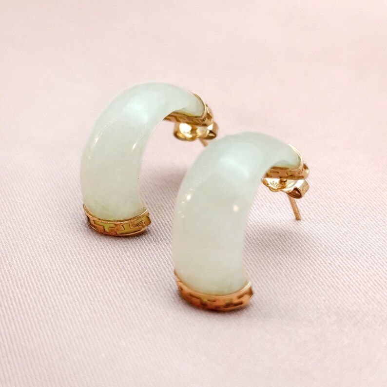 May include: A pair of jade hoop earrings with gold accents. The earrings have a curved, crescent shape with a pale green hue. The gold detailing is visible at the top and bottom of the jade hoops. The earrings are displayed on a pink surface.