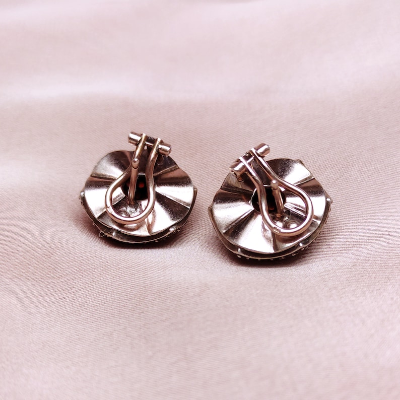May include: A pair of silver tone earrings with a round, scalloped design. Each earring features a red gemstone in the centre.