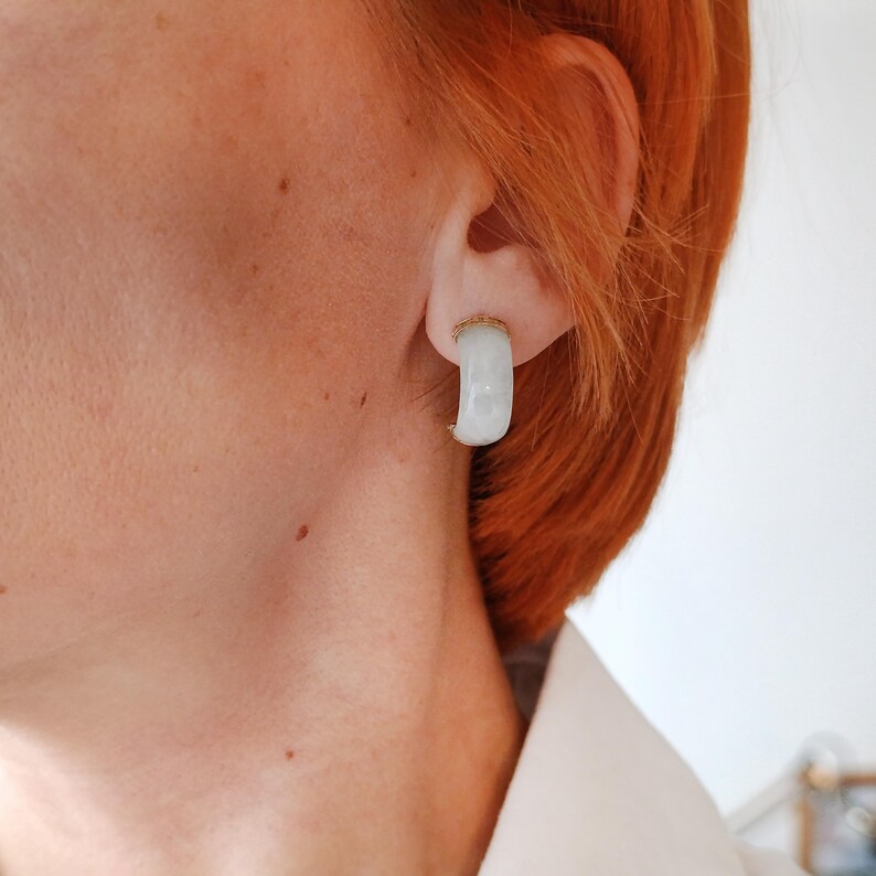 May include: A close-up of a white and gold earring. The earring is a small, curved hoop with a gold band at the top. The white portion of the earring has a marbled appearance. The earring is in the ear of a person with red hair.
