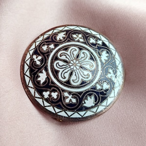 May include: A round, silver-toned brooch with a white and black enamel design. The design features a central flower with intricate details and a surrounding border of leaves and swirls.