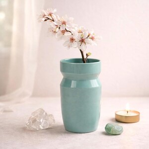 Puede incluir: Un jarrón de cerámica de color verde azulado claro con una rama de delicadas flores blancas y rosas. Un cristal transparente, una piedra verde y una vela dorada encendida están cerca. El jarrón es cilíndrico.