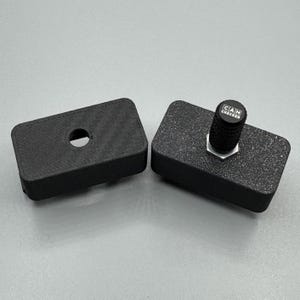 Puede incluir: Dos componentes rectangulares negros. Uno tiene un agujero central, el otro tiene una superficie texturizada y un botón con el texto "CAN CHECKED". Probablemente son partes de un dispositivo o sistema más grande.