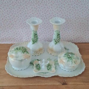 May include: A white porcelain vanity set featuring two candlesticks, two lidded jars, and a small central dish, all arranged on a matching scalloped tray. Each piece is decorated with hand-painted green lily of the valley floral motifs.