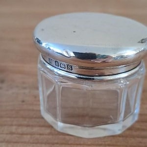 May include: A small, clear glass jar with a faceted, octagonal body and a polished silver lid. The silver lid features a series of small, stamped hallmarks along its side edge, indicating its metallic composition and origin.