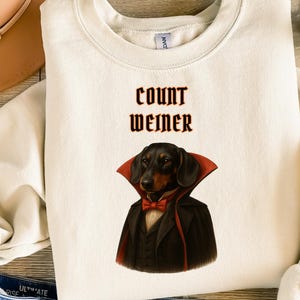 May include: Cream-colored sweatshirt with the text "Count Weimer" in dark red. The graphic features a dachshund dressed as a vampire, wearing a black suit, red cape, and bow tie. A fun Halloween-themed design.