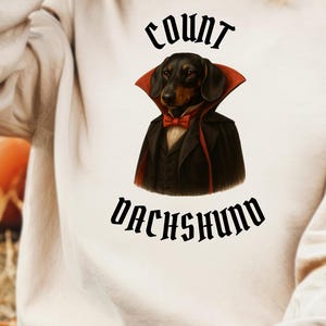 Puede incluir: Sudadera beige con un gráfico de un perro salchicha disfrazado de vampiro, con un traje negro, pajarita roja y capa. El texto "COUNT DACHSHUND" está encima y debajo de la imagen en una fuente gótica.