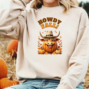 Pode incluir: Uma camisa de moletom branca com o texto "Howdy Fall!" e uma ilustração de desenho animado de uma vaca marrom usando um chapéu de cowboy e segurando uma abóbora.