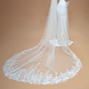 Puede incluir: Vestido de novia blanco con una larga cola y un velo a juego. El velo y la cola están adornados con delicados bordados florales. El vestido es de diseño sencillo y elegante.