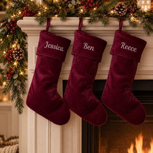 May include: Three deep burgundy velvet Christmas stockings hanging in a row. Each stocking features a folded cuff embroidered with a white name: Jessica, Ben, and Reece. The stockings are suspended from a mantelpiece decorated with greenery.