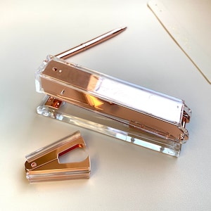 May include: A rose gold stapler and a rose gold staple remover. The stapler is made of clear plastic with a rose gold metal top. The staple remover is also made of clear plastic with a rose gold metal top.
