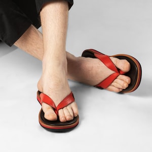 Puede incluir: Una persona con pantalones negros y una camisa negra de manga larga está sentada con las piernas cruzadas. Lleva sandalias de dedo de cuero rojo con suelas marrones.