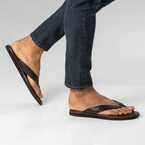 Puede incluir: Un par de sandalias de cuero marrón con tiras, de diseño sencillo. Las sandalias tienen una tira de cuero marrón que va entre los dedos y una tira de cuero marrón que va alrededor del tobillo. Las sandalias las lleva una persona que lleva pantalones vaqueros azules.
